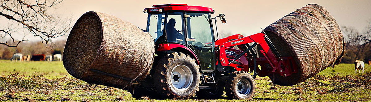 Mahindra® Loader 2545-L lifting two bales of hay in the middle of a cow field
